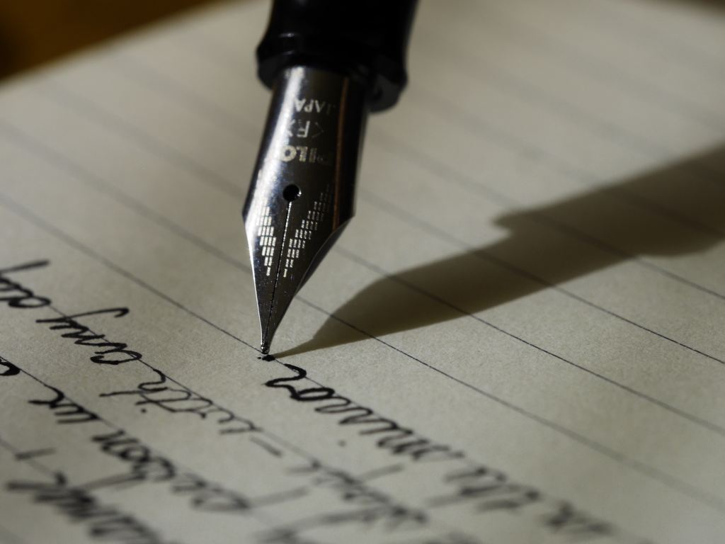 Image of a fountain pen being used to write on cream, lined paper.  Several lines of text are visible, and the pen has just written a full stop.